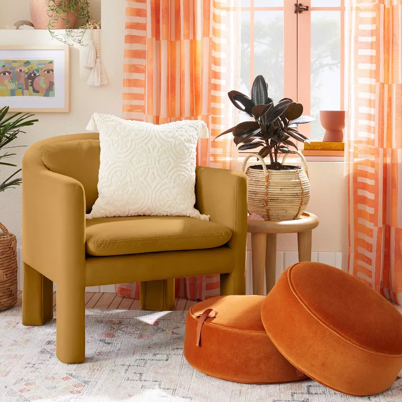 Mustard yellow velvet accent chair with white pillow next to round side table and orange floor cushions in bright living room
