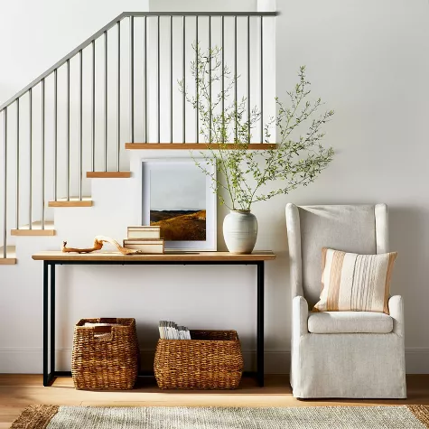 Modern entryway console table with wicker baskets beneath staircase, decorated with books, vase and greenery