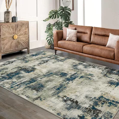 Modern abstract area rug in blue and beige tones displayed in living room with brown leather sofa and wooden cabinet