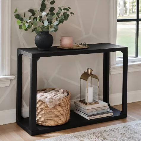 Black console table with two shelves styled with vase, candle, basket, and books in modern entryway