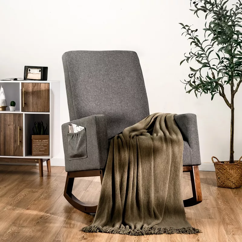 Gray upholstered rocking chair with olive throw blanket in modern nursery with wood flooring and storage unit