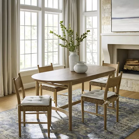 Round wooden dining table with four chairs in bright room featuring large windows, fireplace, and decorative vase centerpiece