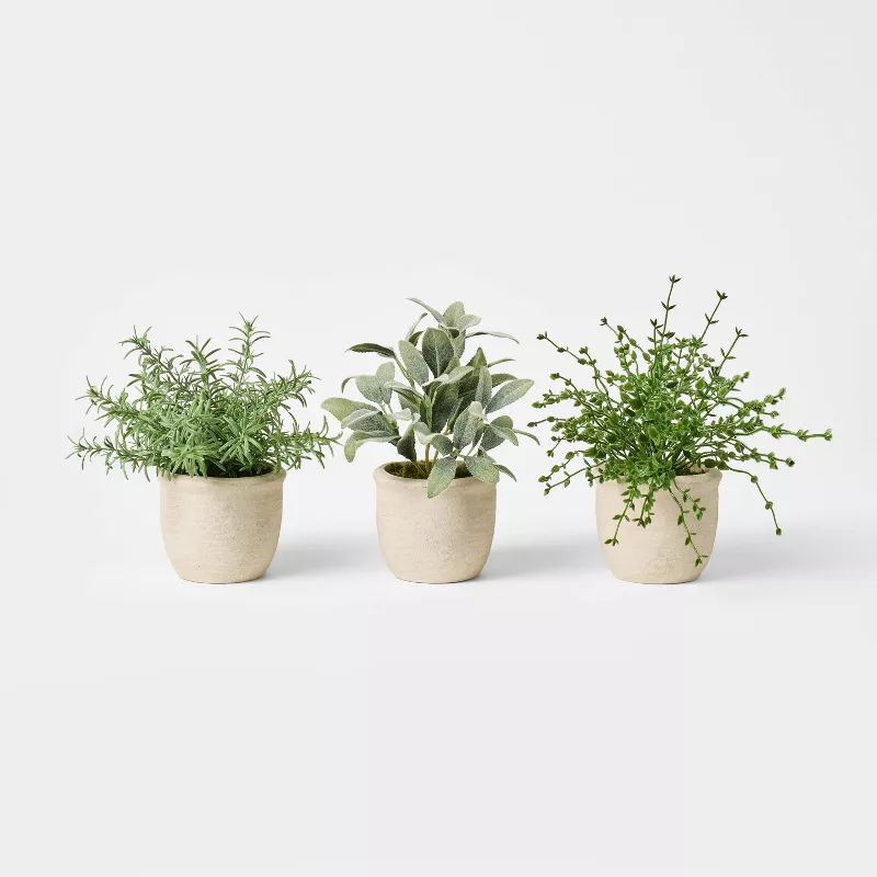 Three realistic faux plants in beige pots including herbs and greenery arranged on white surface