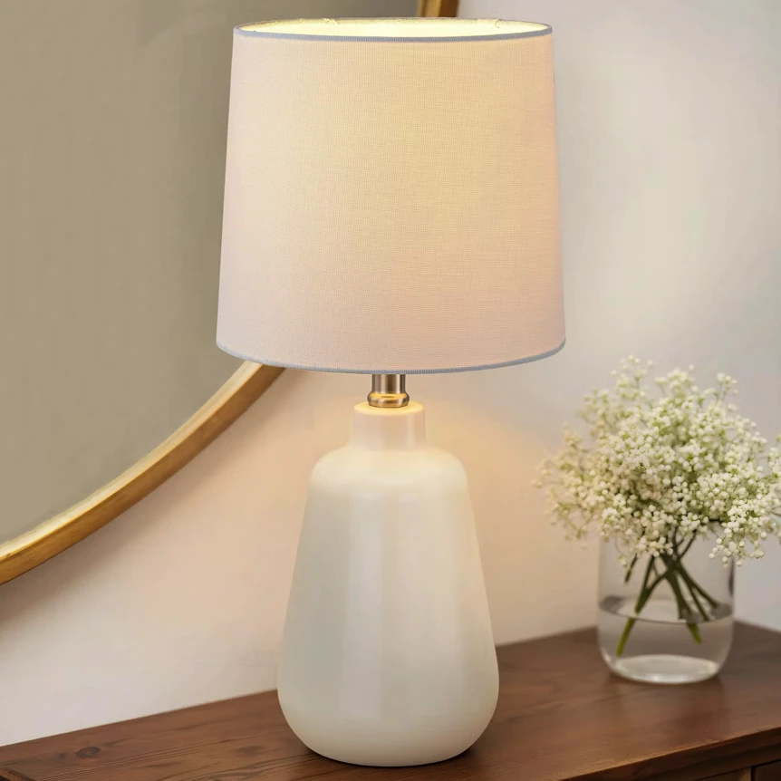 White ceramic table lamp with cream shade on wooden surface next to small vase with flowers
