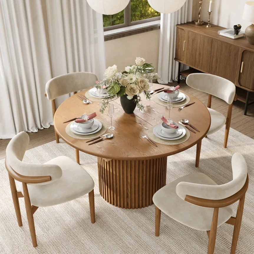 Round wooden dining table with fluted pedestal base and upholstered chairs in small dining room with natural light