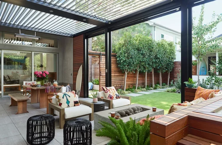 Modern patio with pergola, outdoor furniture, fire pit, and wooden fence surrounded by lush greenery and lawn