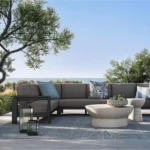 Modern gray sectional sofa with decorative pillows on outdoor patio with blue rug, concrete tables, and ocean view