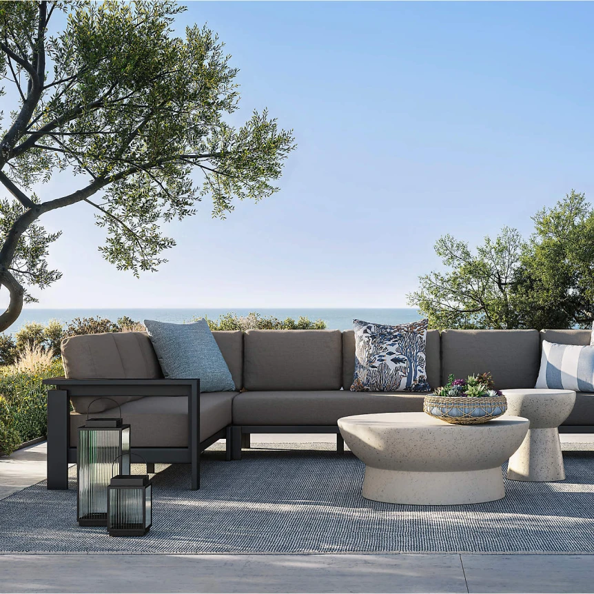 Modern gray sectional sofa with decorative pillows on outdoor patio with blue rug, concrete tables, and ocean view