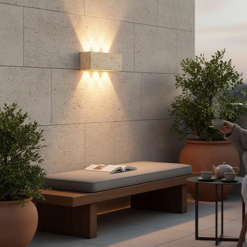 Modern concrete wall sconce with warm LED lighting illuminating minimalist patio seating area with potted plants
