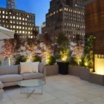 Modern rooftop patio with beige sectional sofa, planters with illuminated trees, and city skyline at dusk