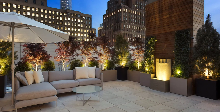 Modern rooftop patio with beige sectional sofa, planters with illuminated trees, and city skyline at dusk