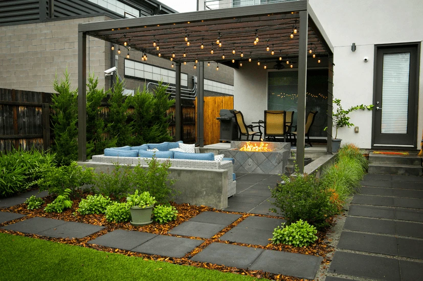 Modern patio with pergola, fire pit, outdoor seating, string lights, and landscaping featuring pavers and greenery