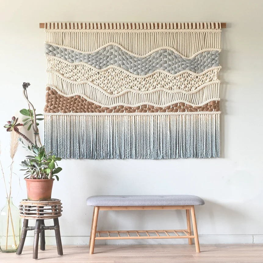 Large macrame wall hanging with geometric patterns and fringe above wooden bench in minimalist living room