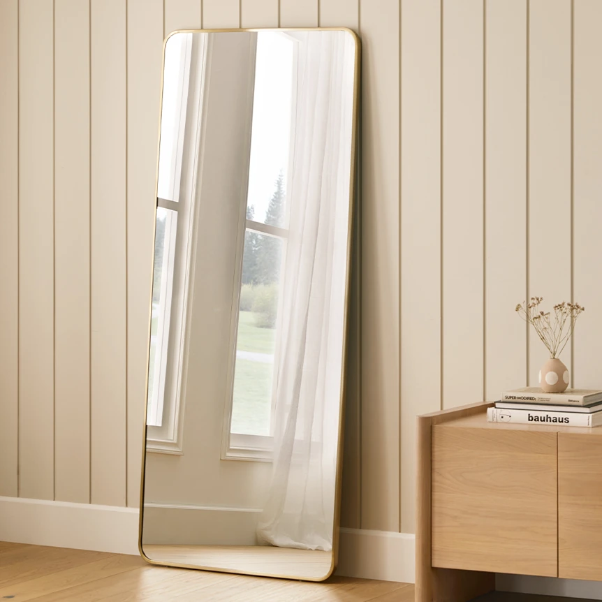 Full-length leaning floor mirror with white frame against beige wood paneled wall next to wooden cabinet