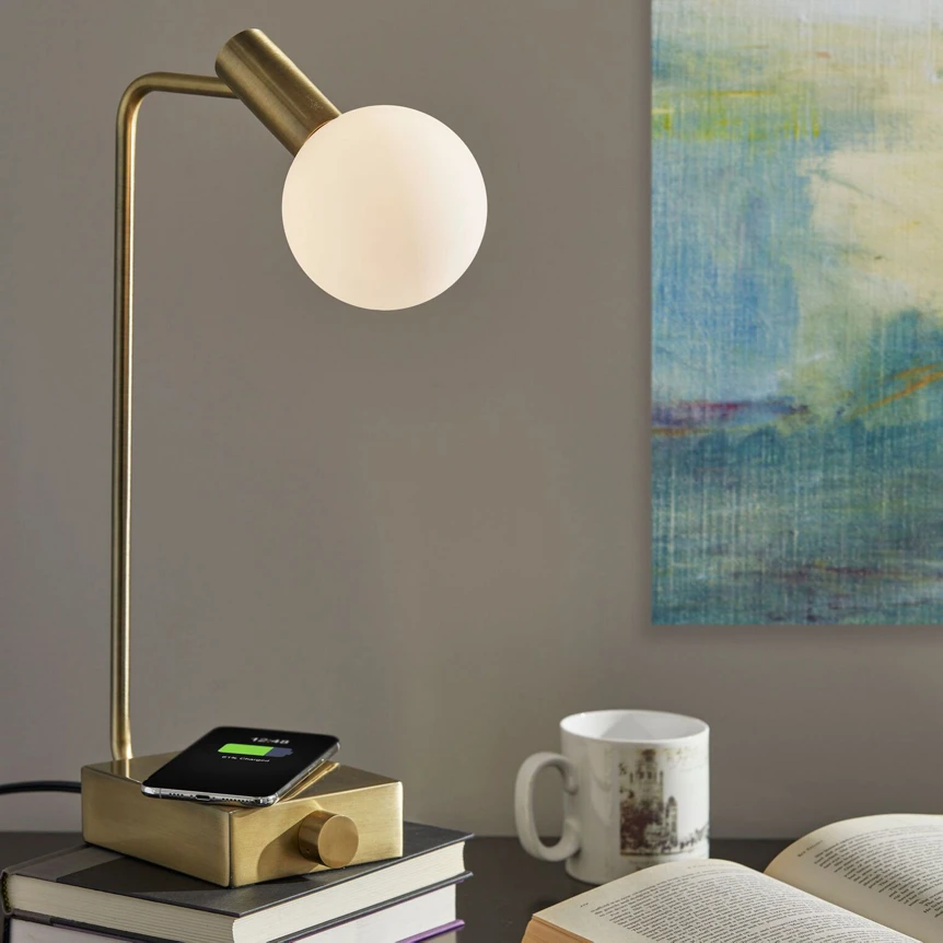 Modern brass desk lamp with globe shade on stacked books beside white mug and abstract wall art