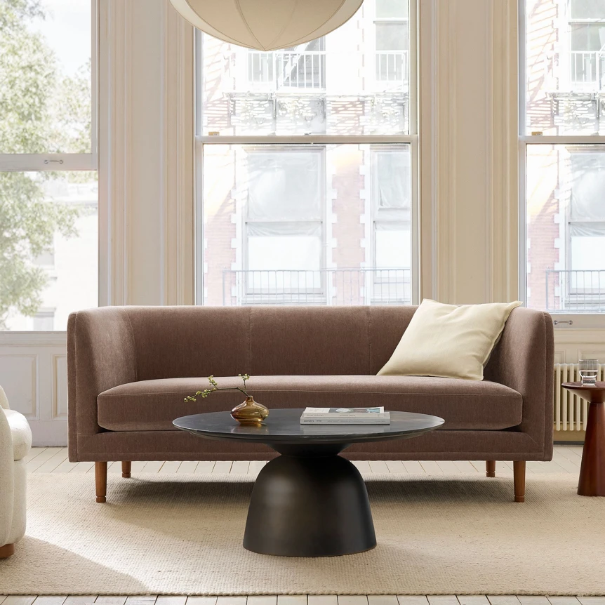Modern brown sofa with wooden legs and round black coffee table in bright living room with large windows