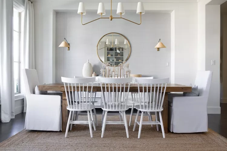 Modern dining room with white Windsor chairs, upholstered bench seating, round mirror, and elegant chandelier