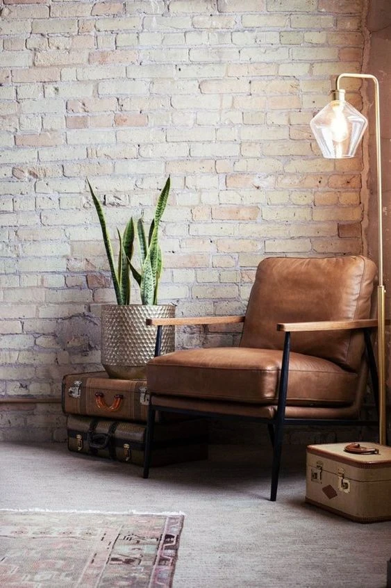 Industrial loft corner with leather chair, floor lamp, exposed brick wall, and snake plant on vintage suitcase table