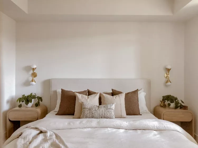 Minimalist bedroom with neutral tones, layered brown pillows, wooden nightstands, and wall sconces in Scandinavian style