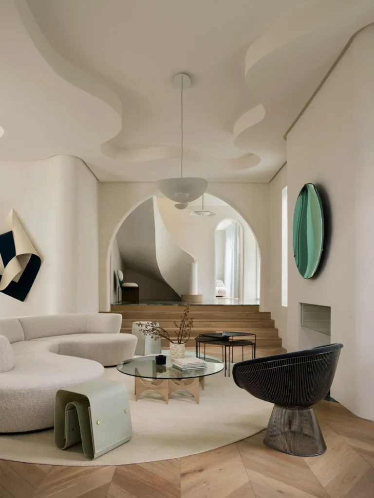 Warm minimalist living room with curved white sofa, natural wood flooring, sculptural lighting, and organic textural elements