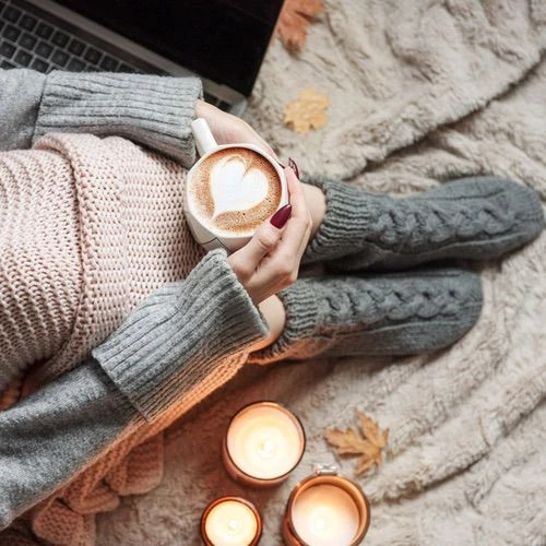 Woman in cozy knit sweater holding latte with heart art surrounded by candles and soft blankets