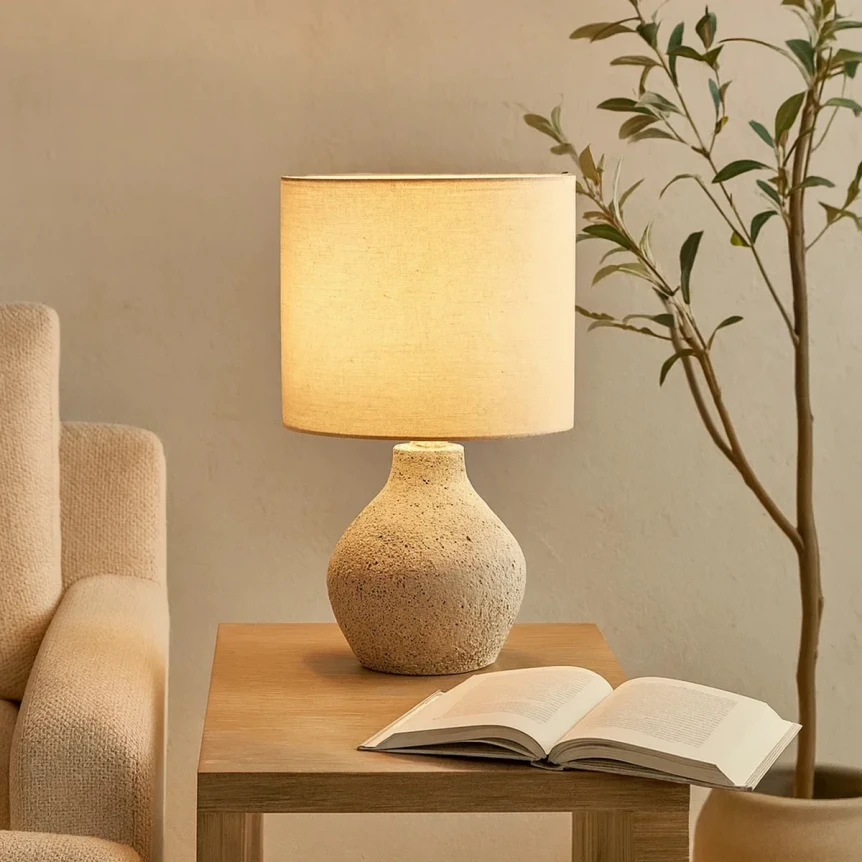 Table lamp with textured beige base and white shade on wooden side table next to open book and plant
