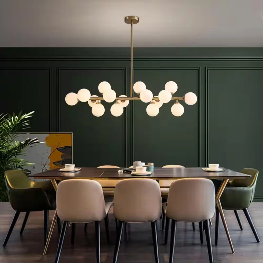 Mid-century modern chandelier with brass finish and globe lights above dining table with velvet chairs against dark green ...