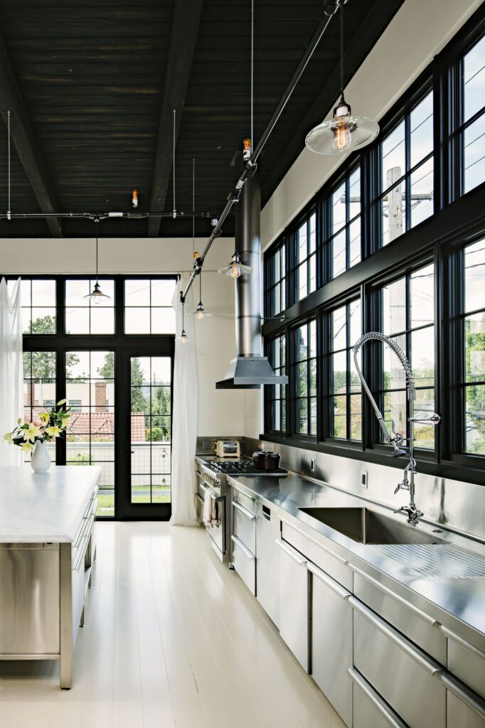 Modern industrial kitchen with floor-to-ceiling black steel windows, white cabinetry, and stainless steel countertops