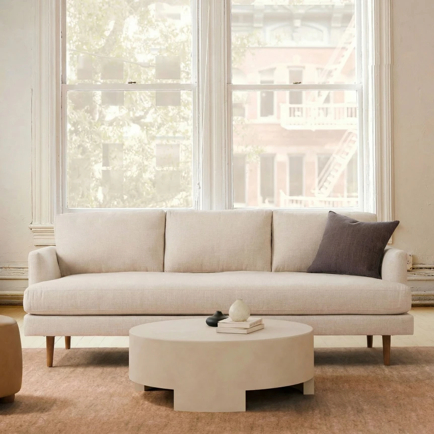 Beige modern sofa with wooden legs and round coffee table in bright living room with large windows