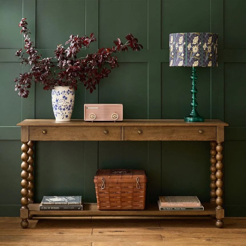 Wooden console table with turned legs against green paneled wall with decorative lamp, vase, and woven basket storage