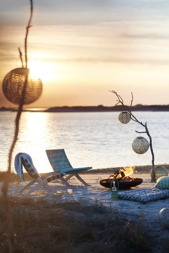 Beach fire pit area with lounge chairs and decorative lanterns at sunset overlooking water