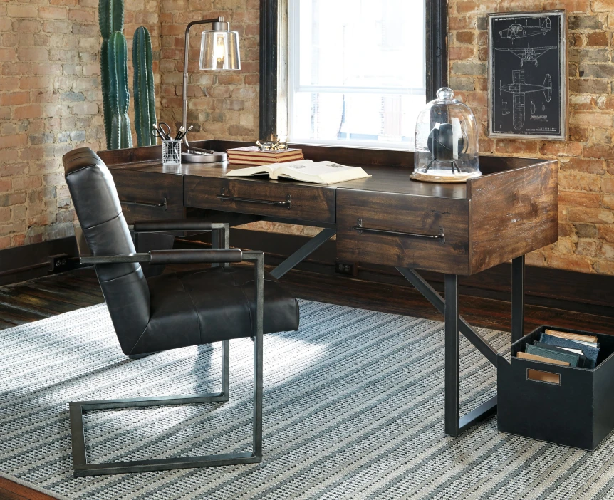 Industrial home office with a wooden desk on black metal legs, gray upholstered task chair, and an exposed brick wall in warm afternoon light