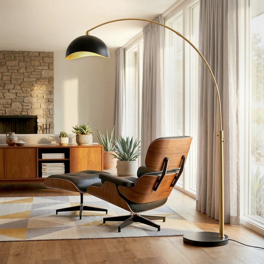 Modern brushed brass arc floor lamp reaching over a tan leather lounge chair next to a home office desk, providing the third lighting layer in warm afternoon daylight