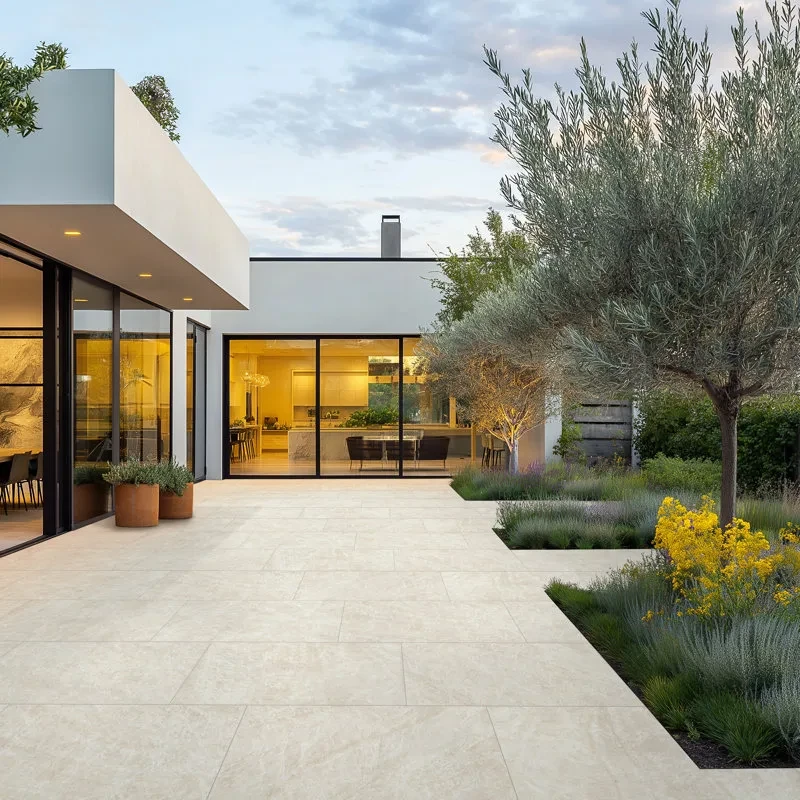 Modern minimalist patio with large glass doors, concrete pavers, and landscaped garden featuring olive tree and natural pl...