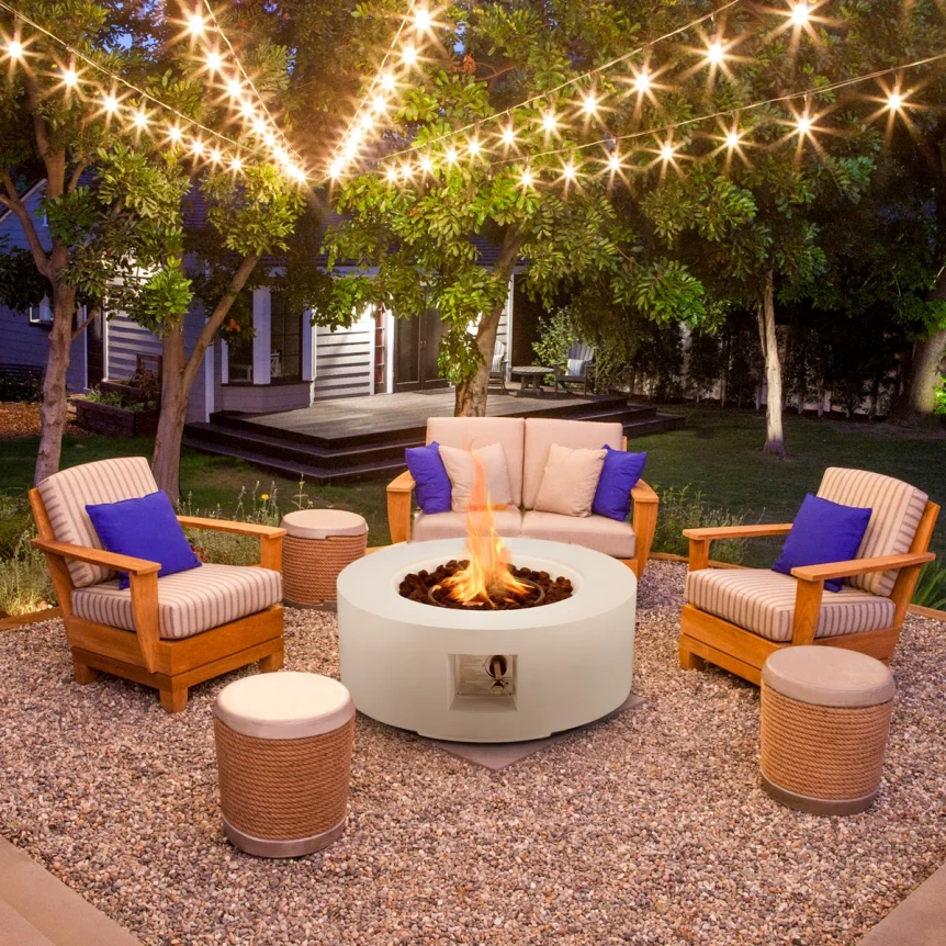 Modern backyard fire pit area at dusk with a circular cast concrete fire pit, low contemporary lounge seating, warm string lights overhead, and pea gravel underfoot