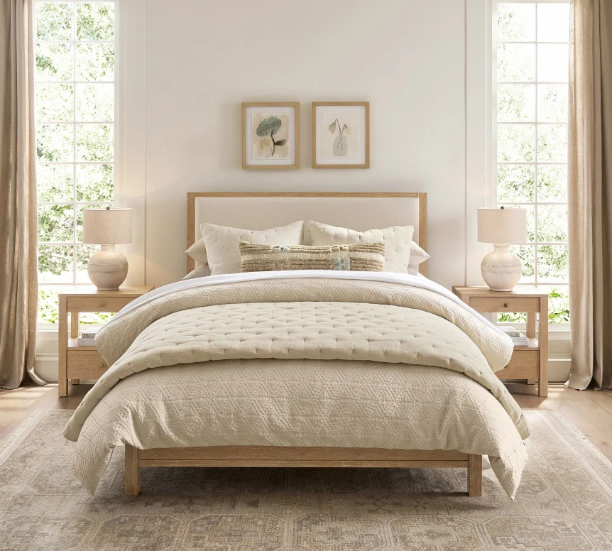 Serene neutral bedroom with wooden bed frame, layered beige bedding, matching nightstands, and natural light from windows
