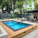 Modern backyard with raised wooden deck pool, outdoor kitchen pavilion, and landscaped garden with patio seating
