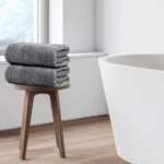 Stack of grey towels on wooden stool next to white bathtub in minimalist modern bathroom
