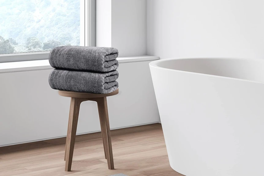 Stack of grey towels on wooden stool next to white bathtub in minimalist modern bathroom