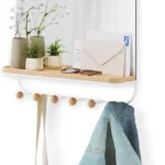 Wall-mounted mirror with wooden shelf and coat hooks holding bags and denim jacket in entryway