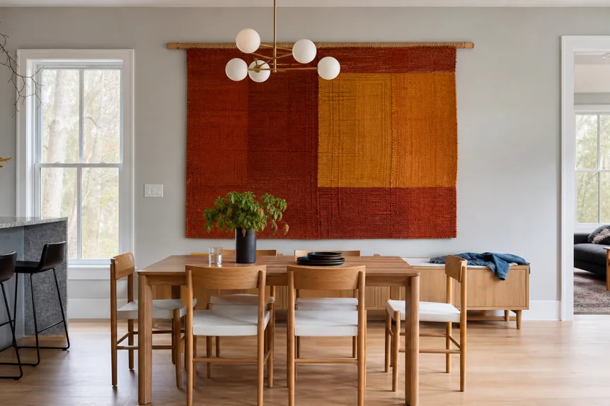 Large woven textile wall hanging in rust and mustard tones in modern dining room with wood furniture and brass chandelier